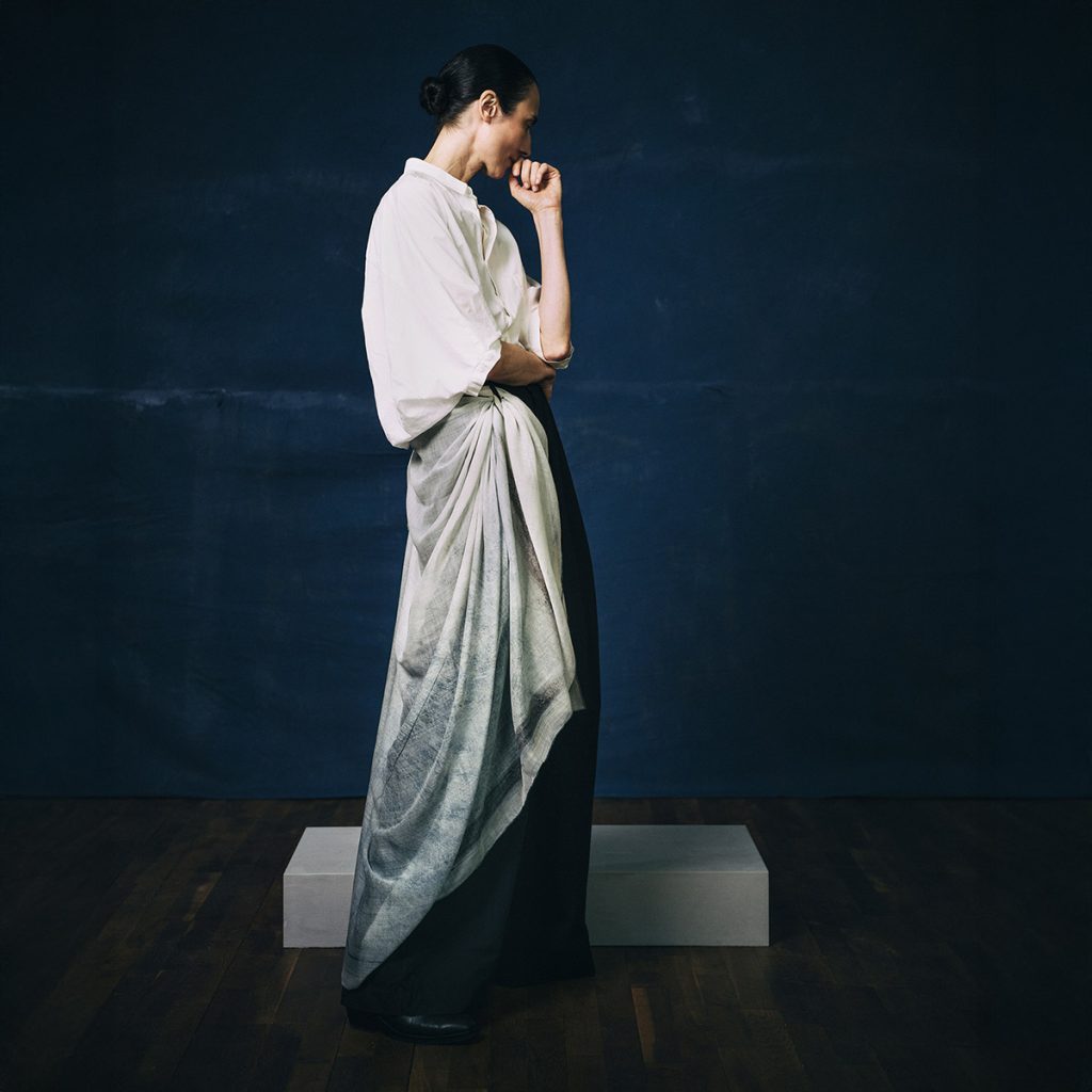 a woman standing in profile against a dark background, she is draped in a pale cashmere scarf from the artist Dot Wade produced by Alba Amicorum