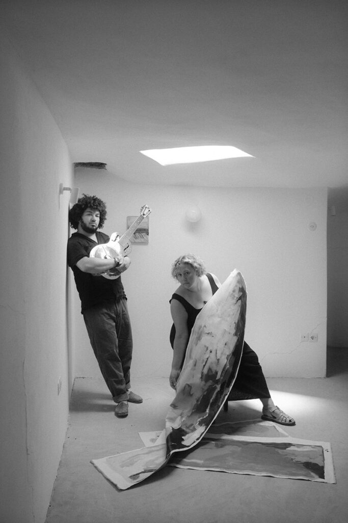 black and white photo of Zuk Zku and Sarah Marlowe Hall in as studio with guitar and painting
